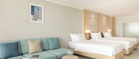 Una habitación de hotel moderna con un sofá de color claro, una mesita auxiliar de mimbre redonda y una cama grande con dos almohadas.