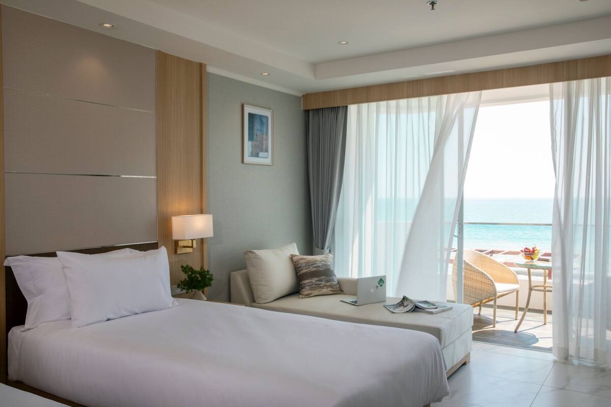 A hotel room with a large bed, a sofa, a laptop, and a view of the sea.