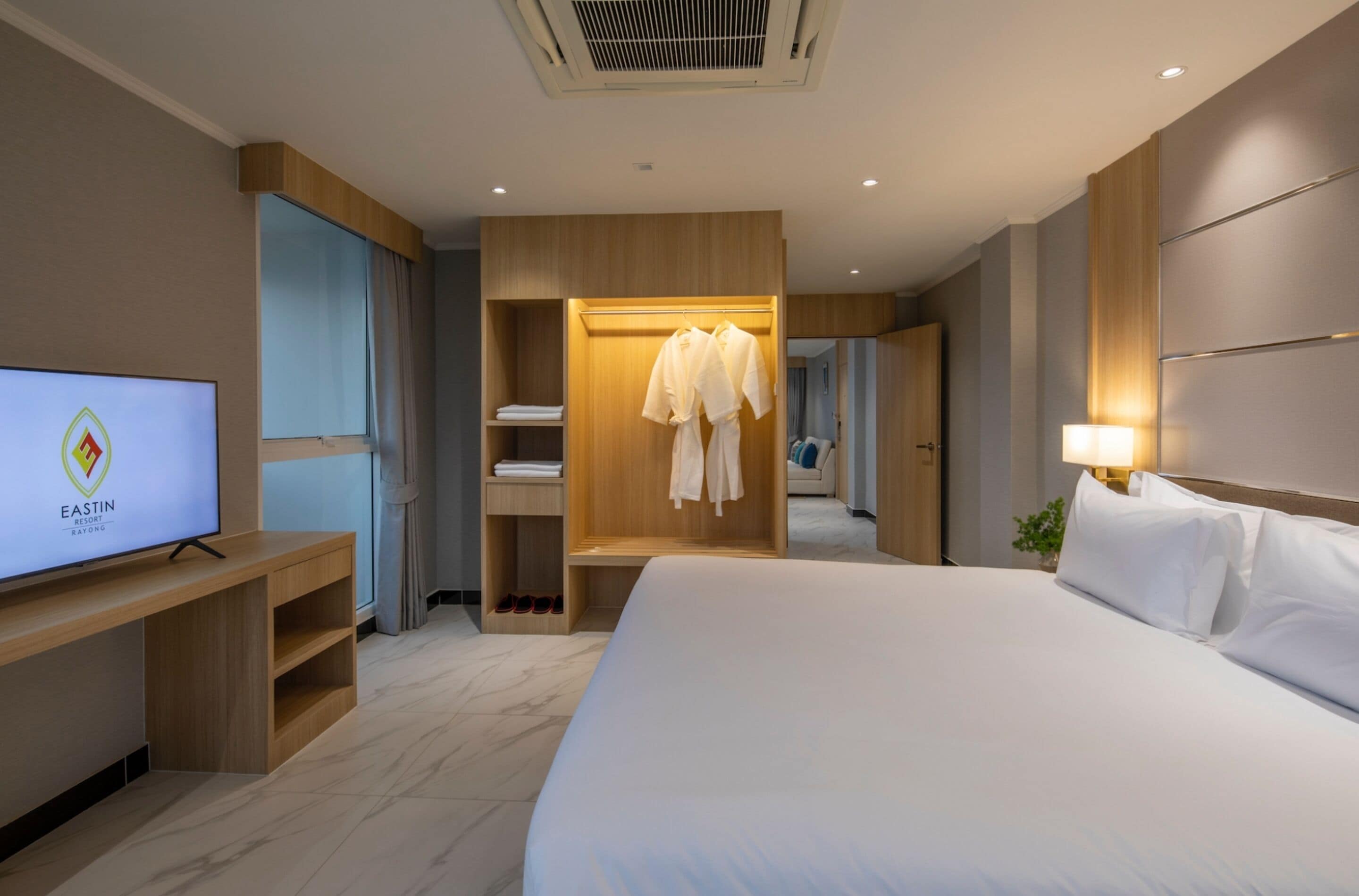 A modern hotel room with a large bed, a TV on a wooden stand, a wardrobe with hanging robes, and a mirror.