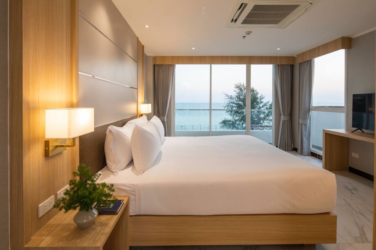 A hotel room with a large bed, a TV on a desk, a plant on the nightstand, and a view of the sea through the window.