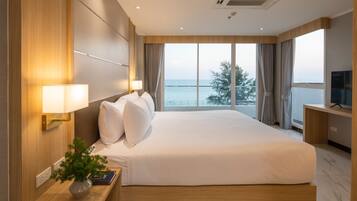 A hotel room with a large bed, a TV on a desk, a plant on the nightstand, and a view of the sea through the window.