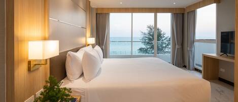 Habitación de hotel con una cama grande, un televisor en un escritorio, una planta en la mesita de noche y vista al mar por la ventana.