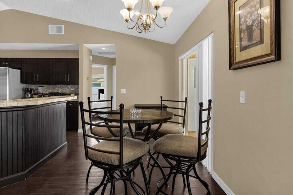 Uma sala de jantar com uma mesa redonda e quatro cadeiras, um candelabro e uma cozinha ao fundo.