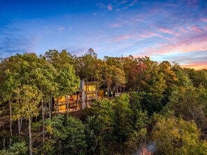 A secluded cabin surrounded by dense forest, featuring a prominent central structure with a visible deck and multiple windows. - Brevard's Cedar Mountain Retreat | 4BR Mountain Views | Hot Tub + Sauna | Near DuPont (Brevard)