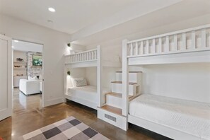 A room with bunk beds, a kitchen area in the background, and a checkered rug on the floor. - Brevard's Cedar Mountain Retreat | 4BR Mountain Views | Hot Tub + Sauna | Near DuPont (Brevard)