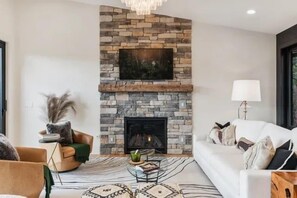 A modern living room with a stone fireplace, a sofa, a coffee table, and a lamp. - Brevard's Cedar Mountain Retreat | 4BR Mountain Views | Hot Tub + Sauna | Near DuPont (Brevard)