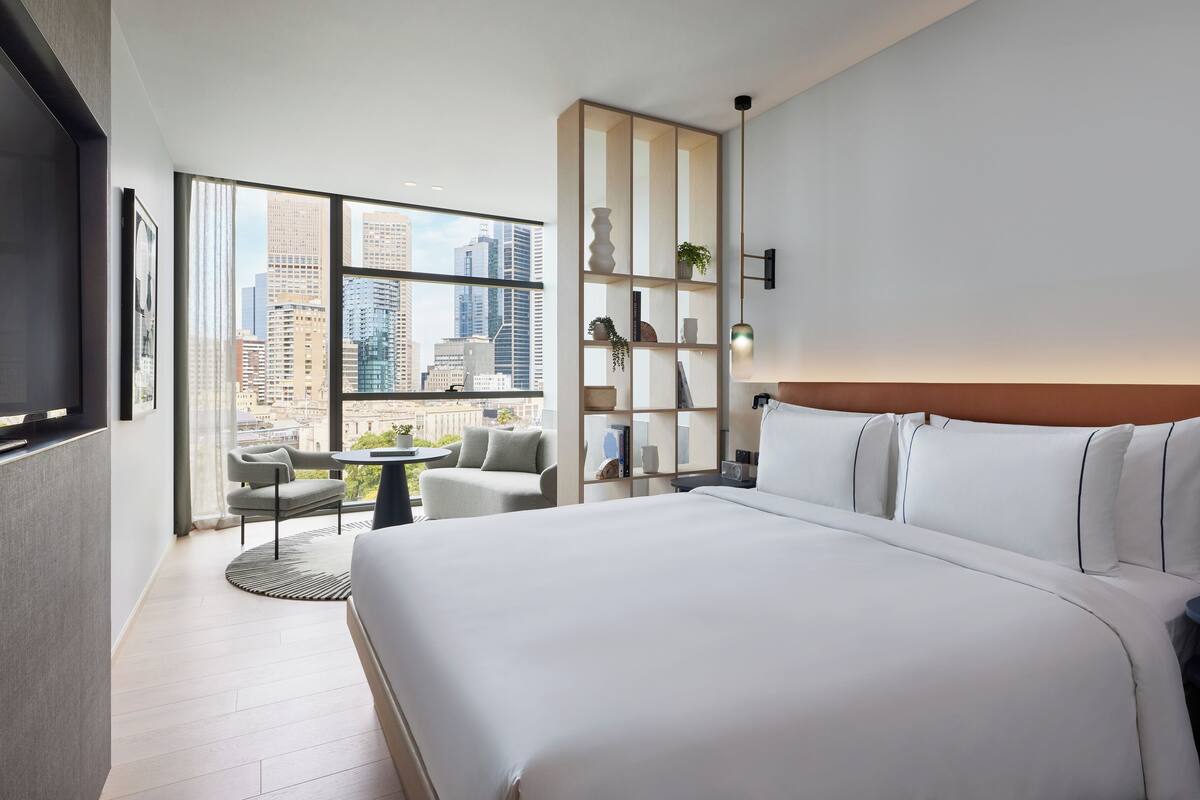 Ein modernes Hotelzimmer mit einem großen Bett, einer Sitzecke mit kleinem Tisch, einer Bücherwand und Blick auf die Stadt durch große Fenster.