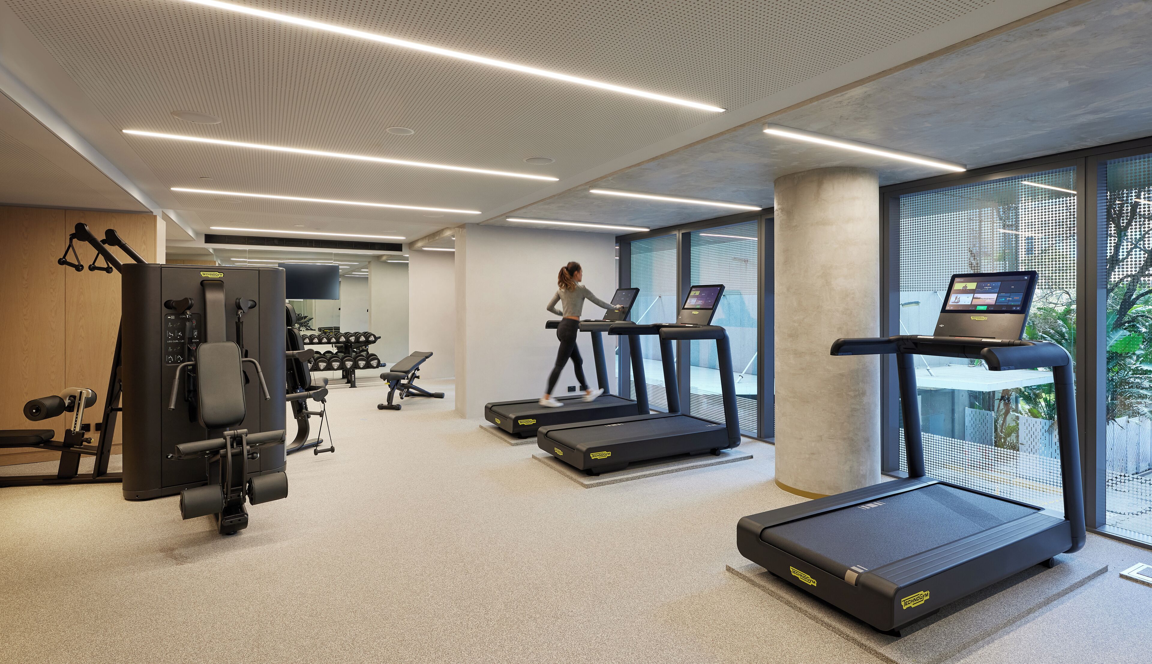 A modern gym with treadmills, weight machines, and a large window offering a view of the outdoors.