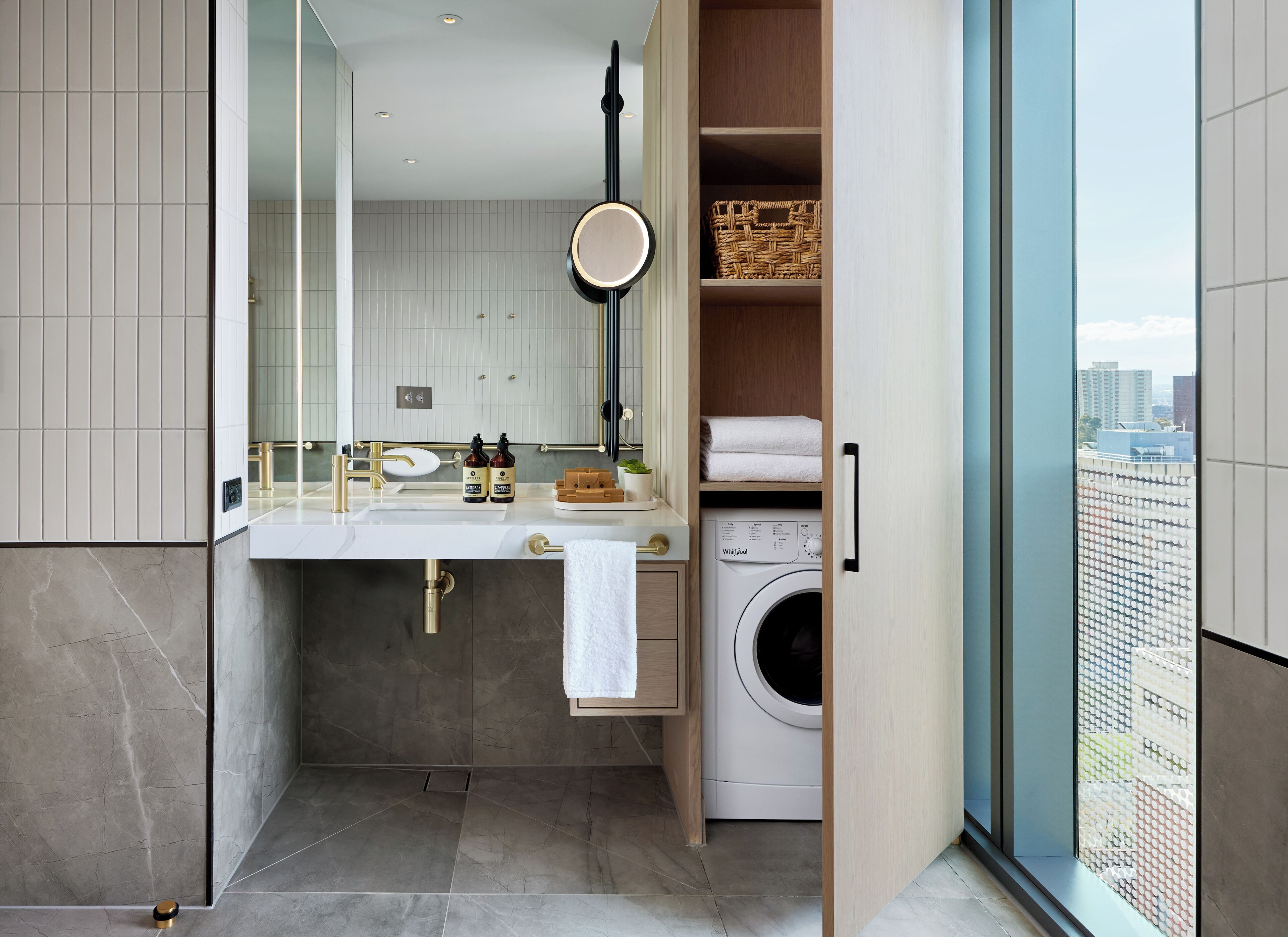 A modern bathroom with a built-in washer and dryer, a large mirror, and a view of the city through a glass door.