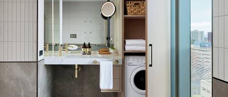 A modern bathroom with a built-in washer and dryer, a large mirror, and a view of the city through a glass door.
