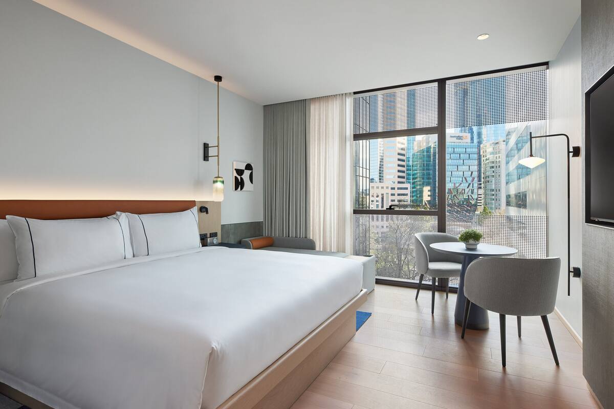 Ein modernes Hotelzimmer mit einem großen Bett, einer kleinen Sitzecke und Blick auf die Stadt durch große Fenster.