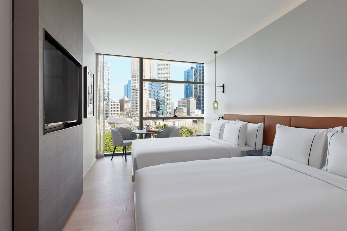 Ein modernes Hotelzimmer mit zwei Betten, einem großen Fenster mit Blick auf die Stadt, einem wandmontierten Flachbildfernseher und einem kleinen Tisch mit einem Stuhl.