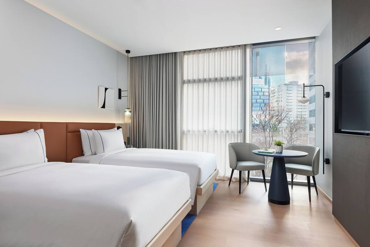 Ein Hotelzimmer mit zwei Betten, einem kleinen Tisch mit Stühlen, einem Fernseher und einem großen Fenster mit Vorhängen.