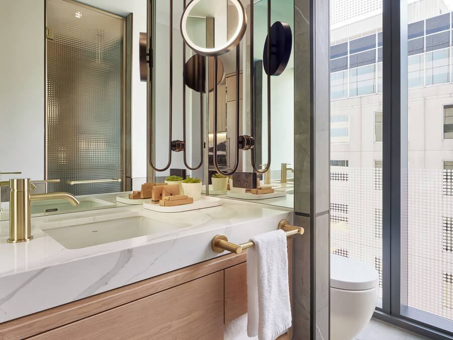 A modern bathroom with a large mirror, a gold faucet, a white marble countertop, and a view of a building through a large window.