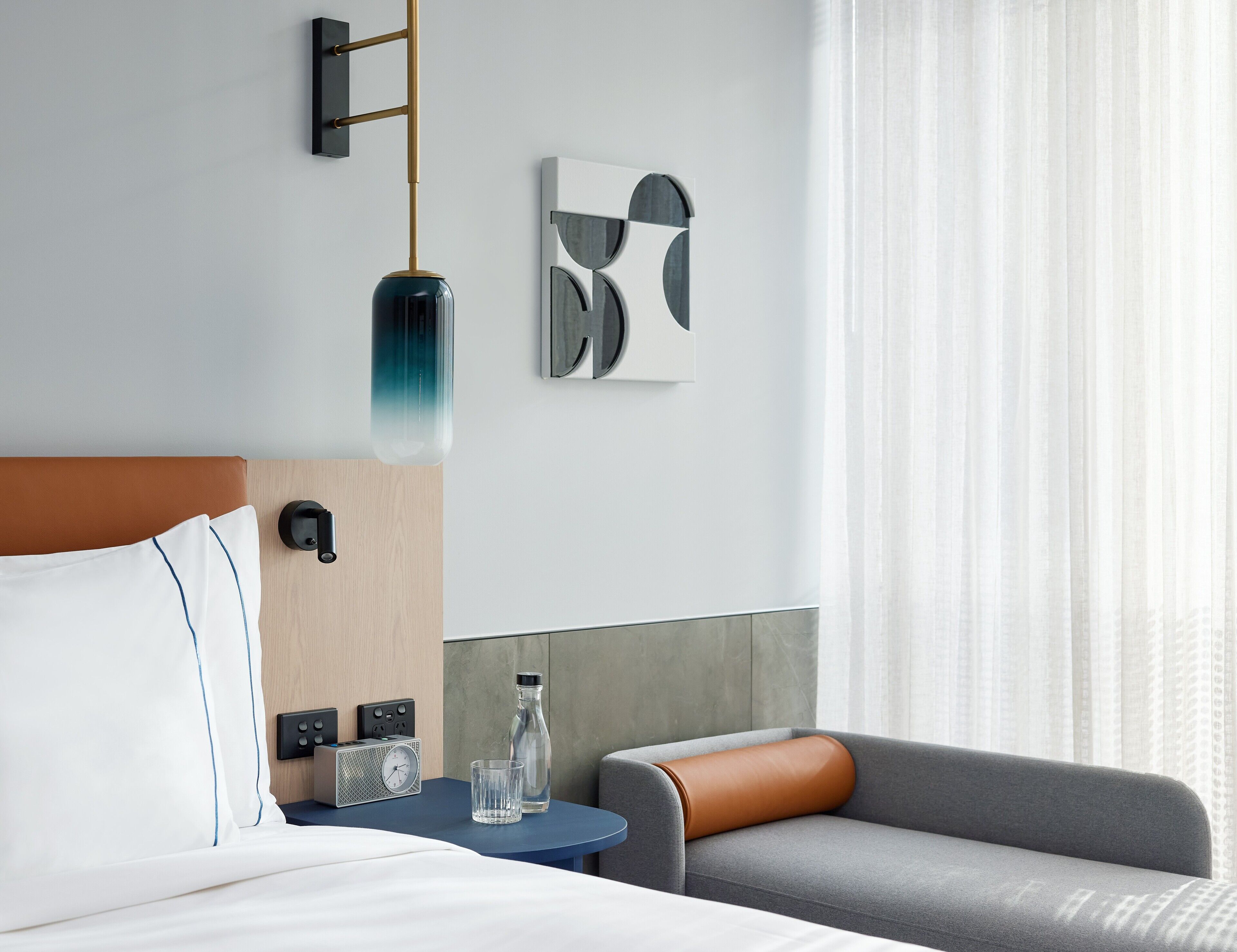A modern hotel room with a bed, a side table with a radio and a bottle, a gray sofa, and a wall-mounted lamp.