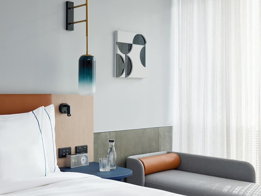 Een moderne hotelkamer met een bed, een bijzettafeltje met een radio en een fles, een grijze bank en een wandlamp.