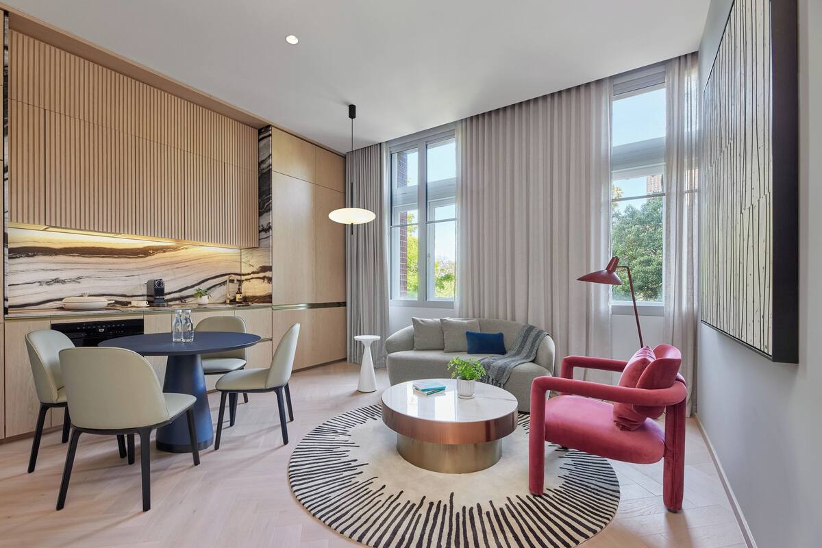 Ein modernes Wohnzimmer mit rundem Couchtisch, einem roten Sessel und einer Couch. Es gibt einen Essbereich mit Tisch und Stühlen sowie eine Küchenzeile mit Herd und Schränken.