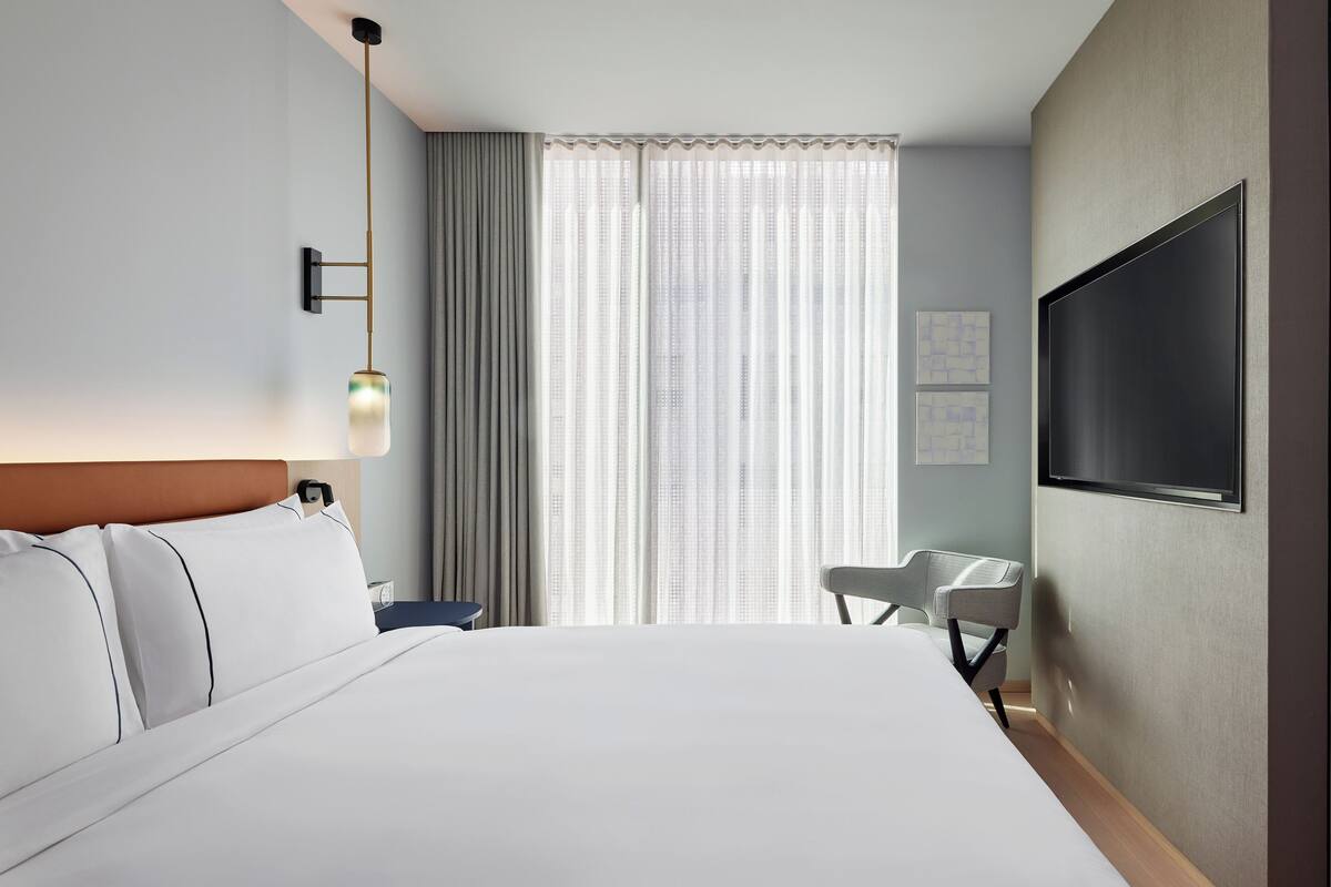 Ein Hotelzimmer mit einem großen Bett, einem Schreibtisch, einem Sessel, einem an der Wand befestigten Fernseher und einem Fenster mit Vorhängen.