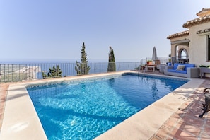 Ein Swimmingpool mit freiem Blick auf das Meer, eine Terrassenanlage mit Gartenmöbeln und ein Gebäude mit einem Ziegeldach.