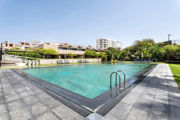 A rectangular swimming pool with a tiled deck, surrounded by greenery and buildings.