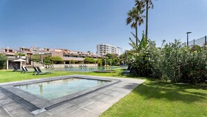 Una piscina amb gandules, una zona enjardinada i un edifici al fons.