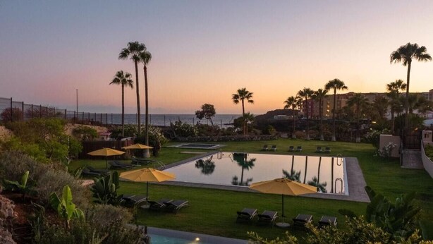 Zona de piscina en un resort con tumbonas, sombrillas y vista al océano al atardecer.