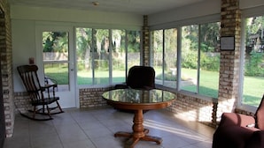 Une terrasse couverte avec une table ronde en verre et des chaises, des murs en briques et de grandes fenêtres donnant sur un jardin.