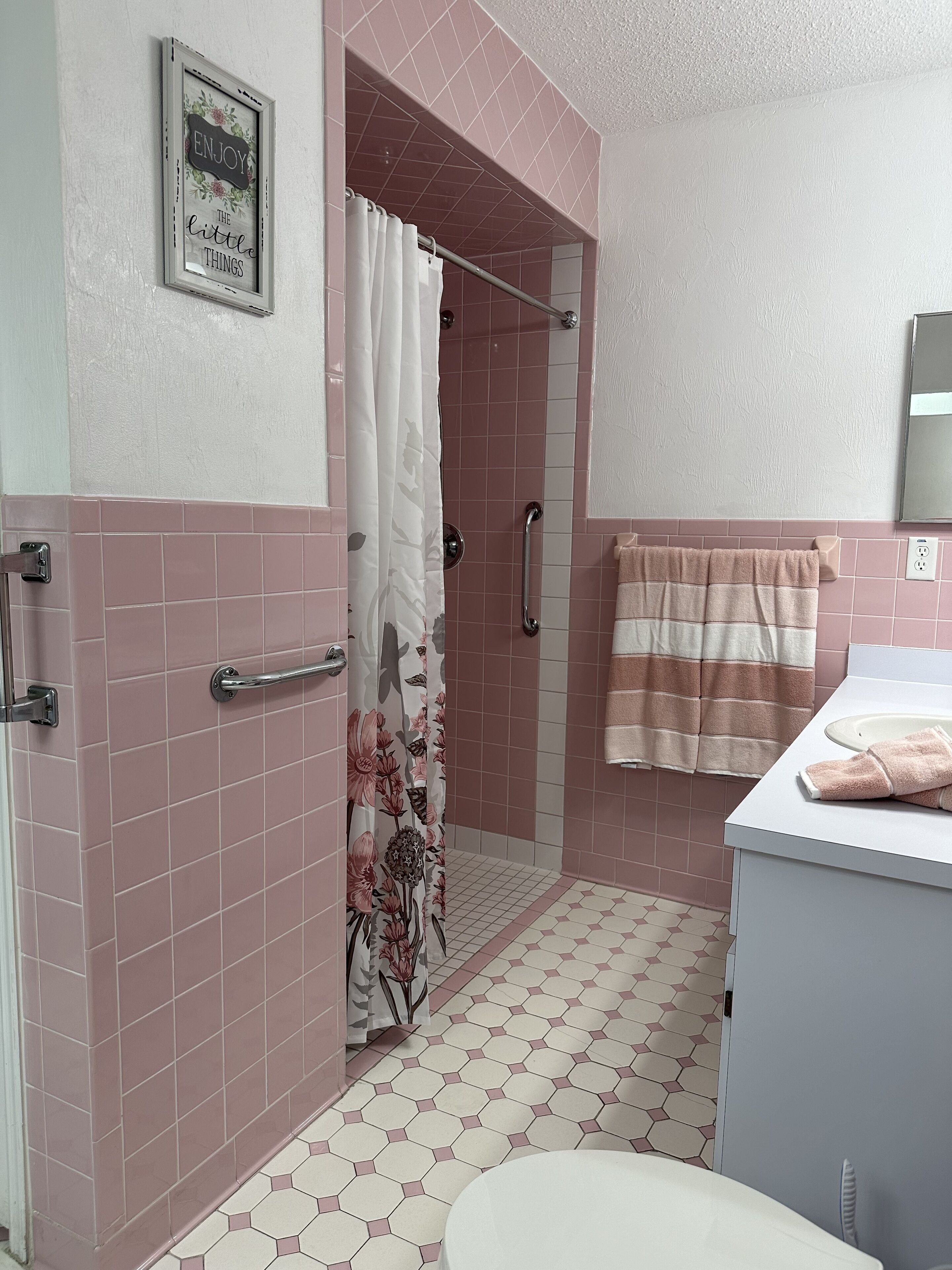 A bathroom with pink tiles, a shower with a floral curtain, a handrail, and a towel rack.