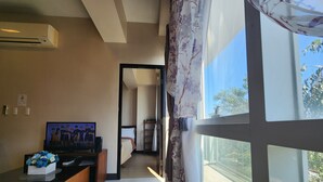 A hotel room with a TV on a wooden stand, a bed with a white headboard, a window with a view of the outdoors, and a ceiling fan. - Your Minimalist One Bedroom Home with Pool with WiFi & Netflix (Lapu-Lapu City)