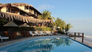 A poolside area with lounge chairs, umbrellas, and a building with a thatched roof.
