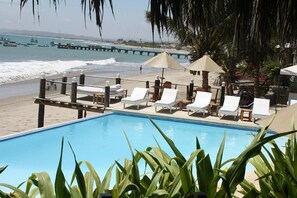 Un área junto a la piscina con sillas de descanso y sombrillas, con vista a una playa y el océano.