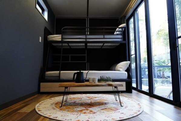 Habitación compacta con literas, una mesita con hervidor, tazas y una planta, y un ventanal con vistas al exterior.