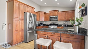 A modern kitchen with wooden cabinets, stainless steel appliances, granite countertops, and two bar stools. - Prime Location, Perfect Stay: Stadium Proximity Home with Pool & Entertainment! (Glendale)