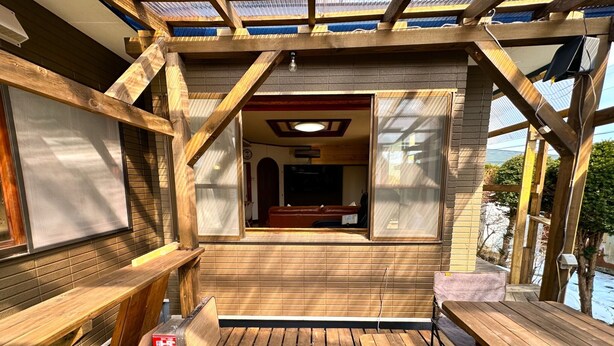 Eine Holzdar Terrasse mit integriertem Sitz eine Tisch, Blick ins Haus durch ein Fenster und eine Metall-Dachkonstruktion.
