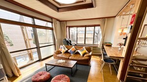 Ein gemütliches Wohnzimmer mit einer braunen Ledersofa, einem Holztisch und einem Essbereich mit Holztisch und Stühlen. Große Fenster sorgen für viel natürliches Licht und bieten einen Ausblick nach draußen.
