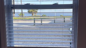 A window with horizontal blinds, a view of a parking area, and a body of water in the distance. - Dolphin View House #2 (Eastpoint)