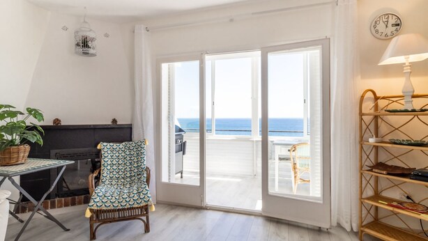 A room with a large sliding glass door leading to a balcony with a view of the sea. There is a wicker chair, a small table, and a shelf with books and a lamp. - Fener de Dalt 90 - Apartment with spectacular view (Llança)
