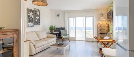 Un salon spacieux avec un canapé, une table basse et deux fauteuils en osier. Une grande fenêtre offre une vue sur la mer.