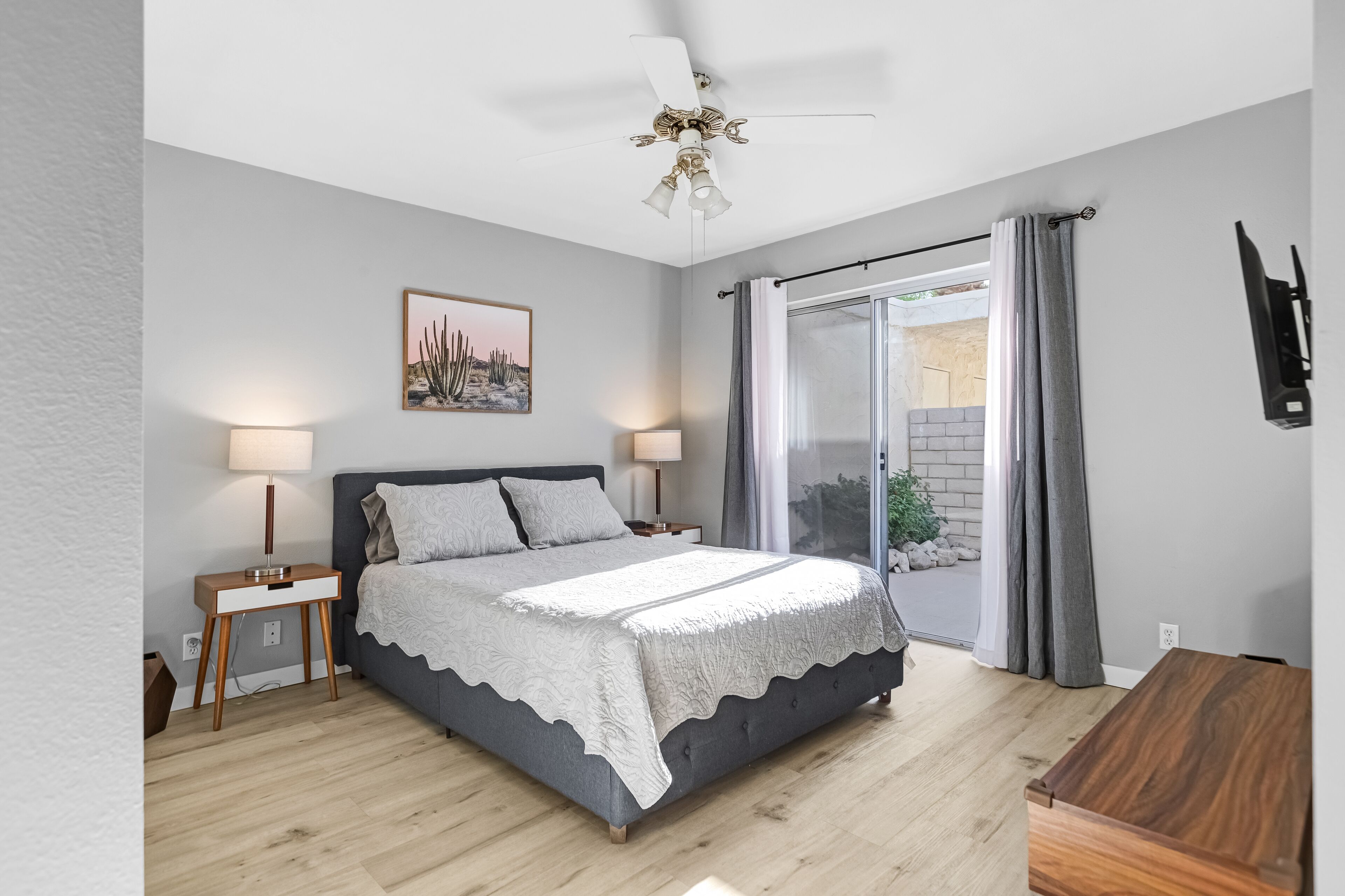 A bedroom with a bed, bedside lamps, a ceiling fan, a window with curtains, and a wall-mounted TV.