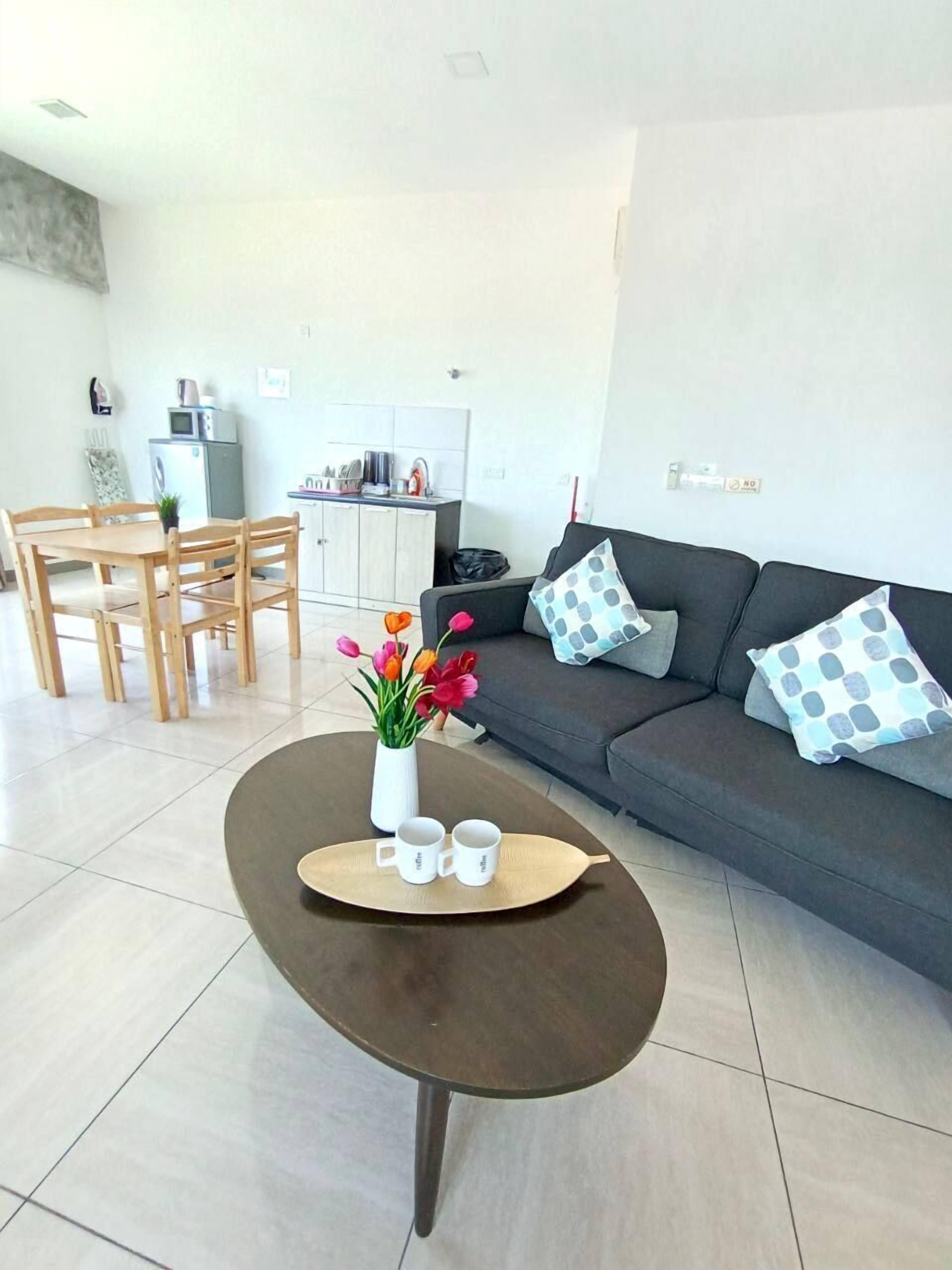 A modern living room with a dark brown coffee table, a grey sofa with patterned cushions, and a dining area with wooden chairs and a small kitchen in the background.