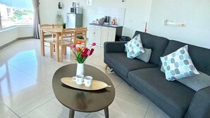 A modern living room with a dark grey sofa, a round coffee table with a vase of flowers, and a dining area with wooden chairs and a small kitchen in the background.