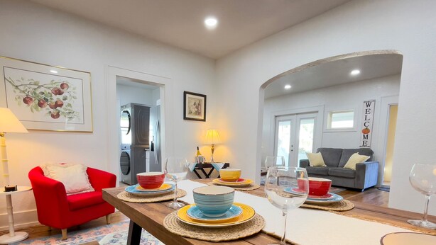 Un coin repas avec une table en bois dressée pour quatre personnes, entourée d’assiettes et de bols colorés, d’un fauteuil rouge et d’un canapé en arrière-plan.