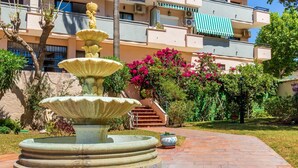 A multi-tiered stone fountain in a courtyard with a tiled border, surrounded by lush greenery and a building with balconies in the background. - Edificio Carihuela - Terrace, Swimming Pool. 300m from the beach (Torremolinos)