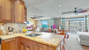 A modern kitchen with wooden cabinets, a marble countertop, and a large window overlooking a balcony with a view of the sea.