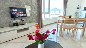 Una habitación de hotel moderna con un televisor de pantalla plana, un rincón de comedor con sillas de madera, una mesita con una bandeja de tazas de café y un florero con flores.