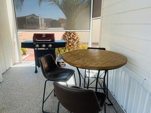 A patio with a round wooden table and two black chairs, a barbecue grill, and a propane tank.