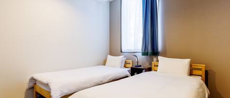A hotel room with two single beds, a window with a blue curtain, a bedside table with a lamp, and a power outlet on the wall.