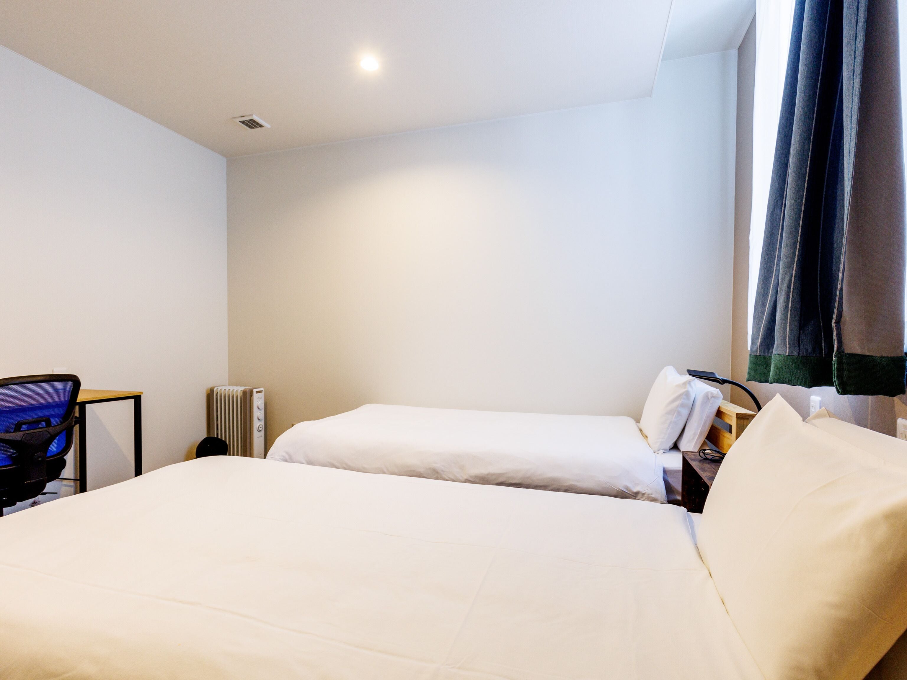 A hotel room with two beds, a desk, a chair, and a radiator.