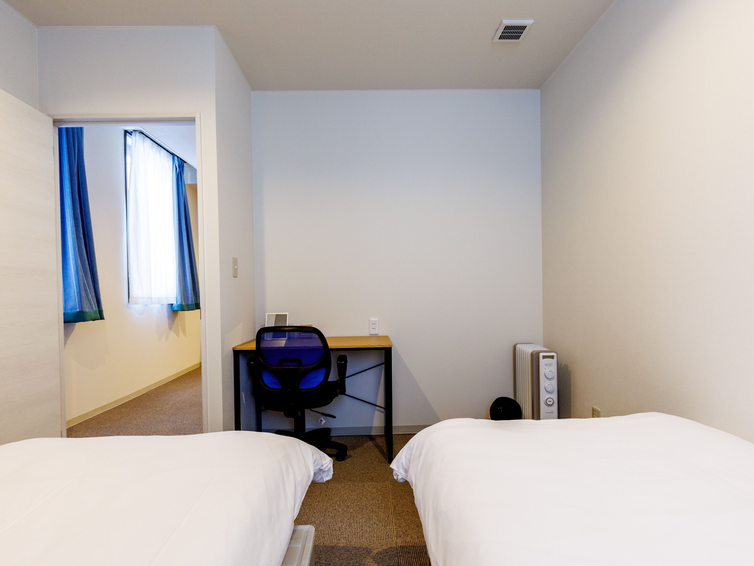 Een hotelkamer met twee bedden, een bureau met stoel, een radiator en een raam met gordijnen.