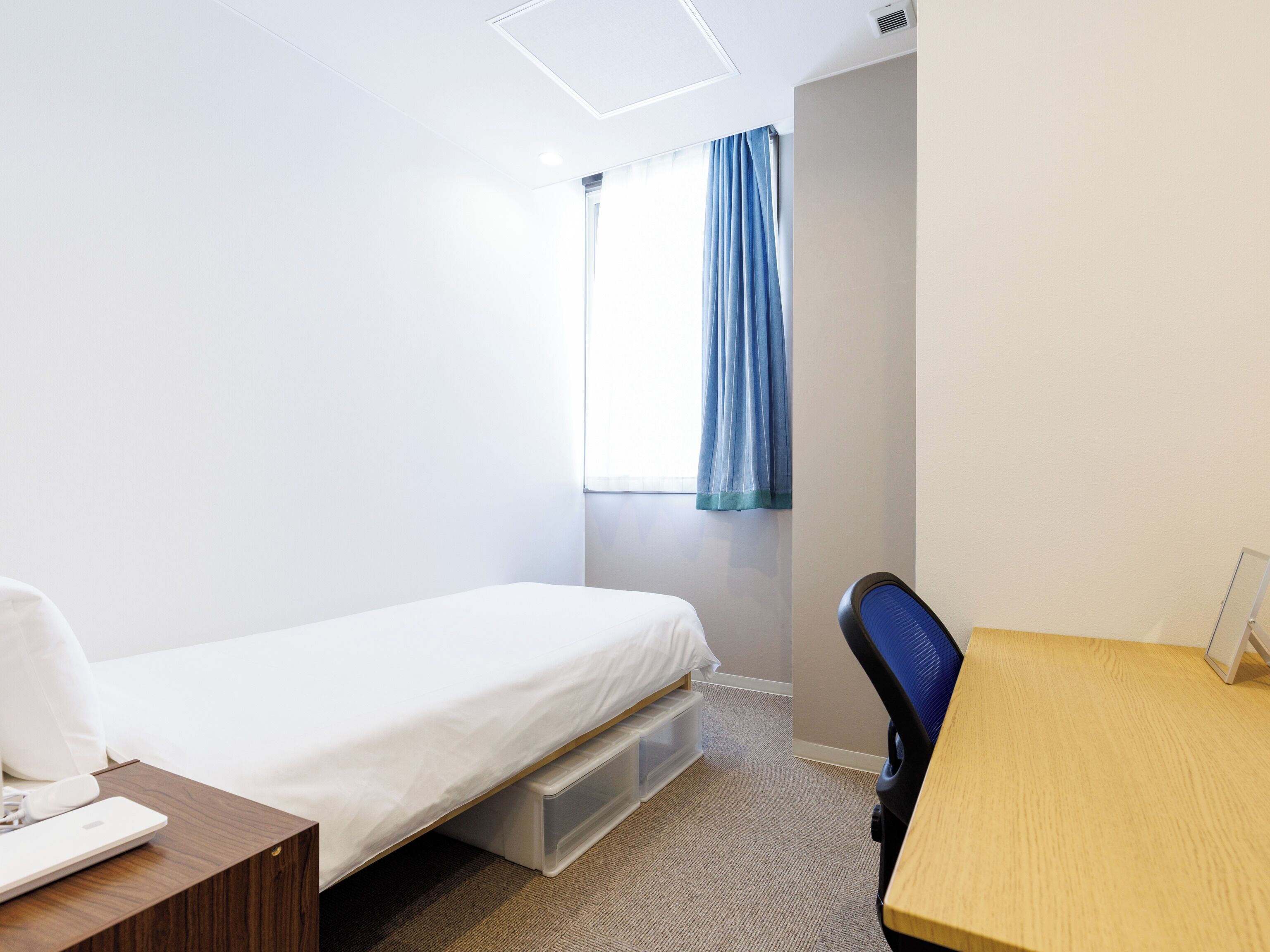 A hotel room with a single bed, a desk with a chair, a window with blue curtains, and a small storage unit under the bed.